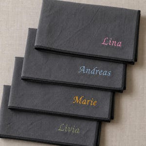 Peut inclure: Quatre serviettes en tissu gris anthracite avec des noms brodés de différentes couleurs. Chaque serviette a une bordure cousue noire et un nom différent brodé en rose, bleu, jaune et vert. Les noms sont Lina, Andreas, Marie et Livia.
