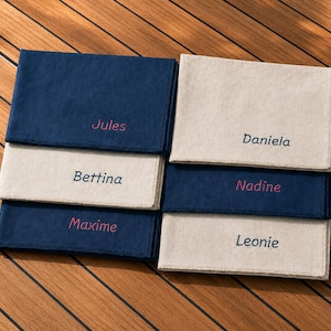 Op de afbeelding: Zes gepersonaliseerde linnen servetten in marineblauw en beige, elk geborduurd met een naam in een contrasterend roze lettertype. De servetten zijn gerangschikt in twee stapels van drie op een houten oppervlak.