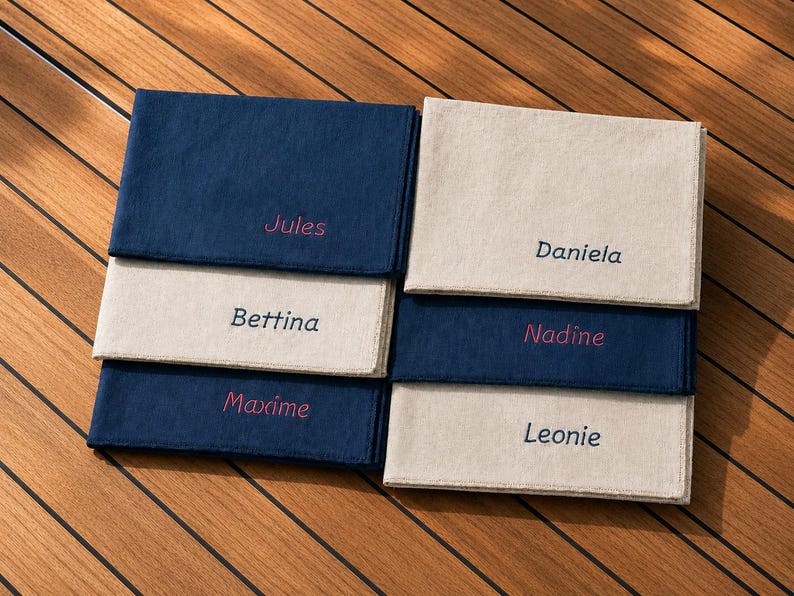 Op de afbeelding: Zes gepersonaliseerde linnen servetten in marineblauw en beige, elk geborduurd met een naam in een contrasterend roze lettertype. De servetten zijn gerangschikt in twee stapels van drie op een houten oppervlak.