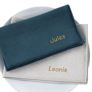 Op de afbeelding: Twee gepersonaliseerde teal en lichtgrijze servetten met geborduurde namen, "Jules" en "Leonie", in goudkleurig garen. De servetten zijn gevouwen en op een wit bord geplaatst.