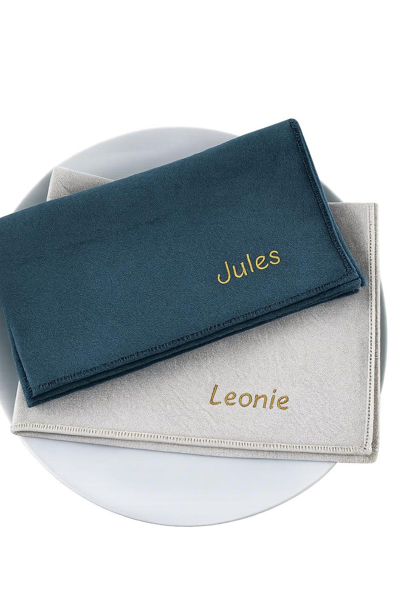 Op de afbeelding: Twee gepersonaliseerde teal en lichtgrijze servetten met geborduurde namen, "Jules" en "Leonie", in goudkleurig garen. De servetten zijn gevouwen en op een wit bord geplaatst.