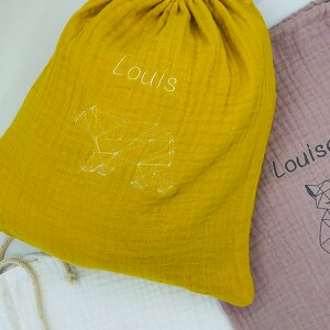 May include: Three drawstring bags made of soft, yellow, pink, and white fabric. Each bag has a geometric animal design embroidered on the front. The yellow bag says "Louis" and has a bear design. The pink bag says "Louise" and has a fox design. The white bag says "rielle" and has a fox design.