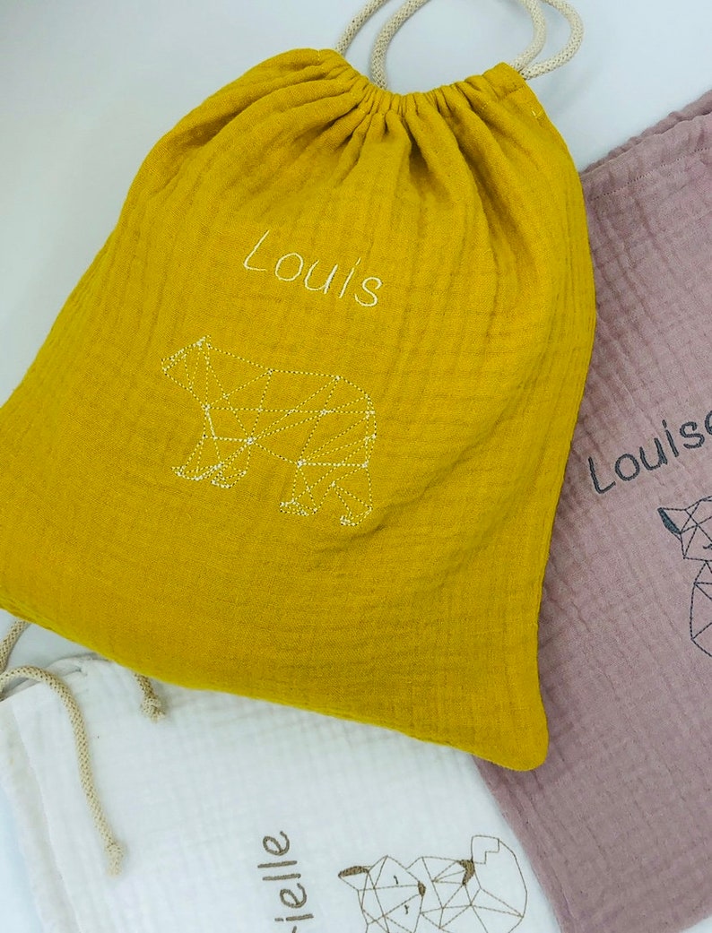 May include: Three drawstring bags made of soft, yellow, pink, and white fabric. Each bag has a geometric animal design embroidered on the front. The yellow bag says "Louis" and has a bear design. The pink bag says "Louise" and has a fox design. The white bag says "rielle" and has a fox design.