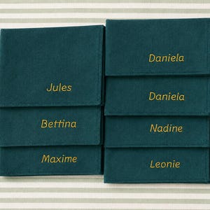 Op de afbeelding: Teal vilten naambordjes met goud geborduurde namen: Jules, Bettina, Maxime, Daniela, Nadine en Leonie. De naambordjes zijn geplaatst op een gestreepte achtergrond.