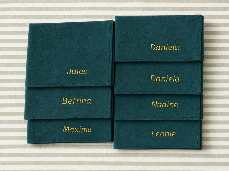 Op de afbeelding: Teal vilten naambordjes met goud geborduurde namen: Jules, Bettina, Maxime, Daniela, Nadine en Leonie. De naambordjes zijn geplaatst op een gestreepte achtergrond.
