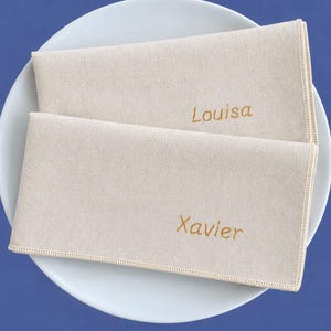 Op de afbeelding: Twee beige linnen servetten met de namen "Louisa" en "Xavier" geborduurd in goudkleurig garen, op een wit bord. De servetten hebben een eenvoudig, elegant ontwerp, perfect voor een formeel diner.