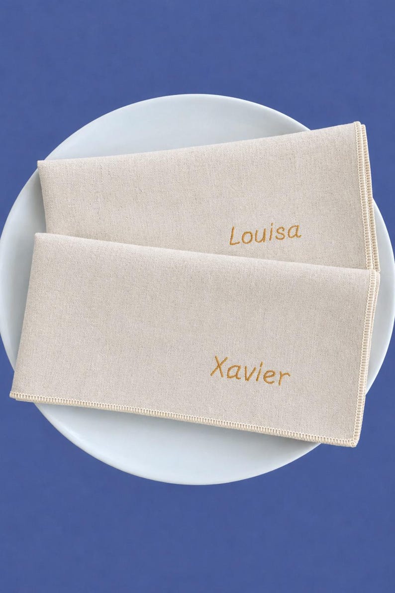 Op de afbeelding: Twee beige linnen servetten met de namen "Louisa" en "Xavier" geborduurd in goudkleurig garen, op een wit bord. De servetten hebben een eenvoudig, elegant ontwerp, perfect voor een formeel diner.