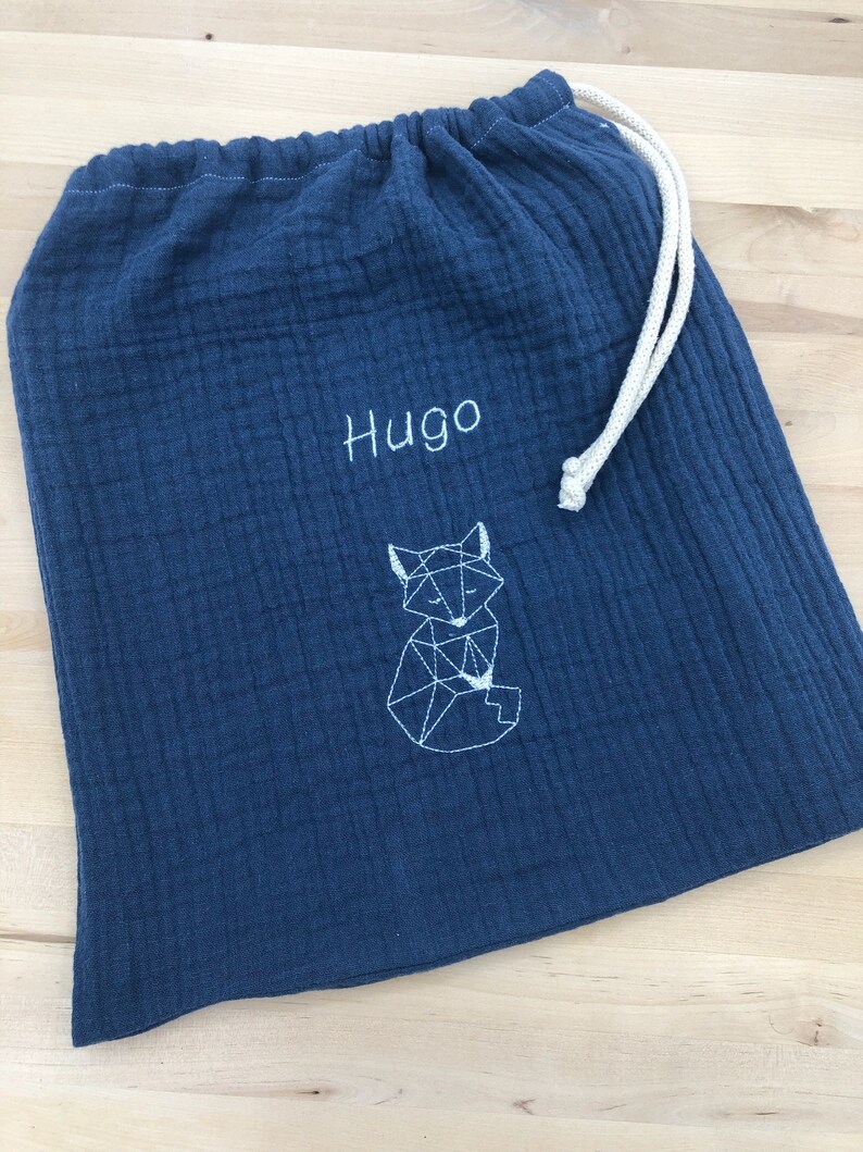 May include: A dark blue fabric drawstring bag with a white geometric fox design and the name "Hugo" embroidered on the front.