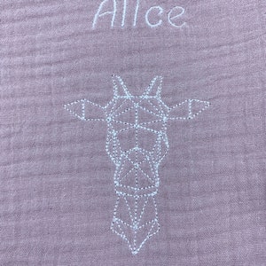 May include: A pale pink muslin baby blanket with white embroidery. The blanket features the name "Alice" and a geometric design of a giraffe.