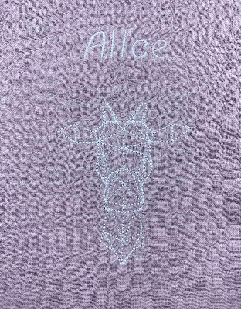 May include: A pale pink muslin baby blanket with white embroidery. The blanket features the name "Alice" and a geometric design of a giraffe.