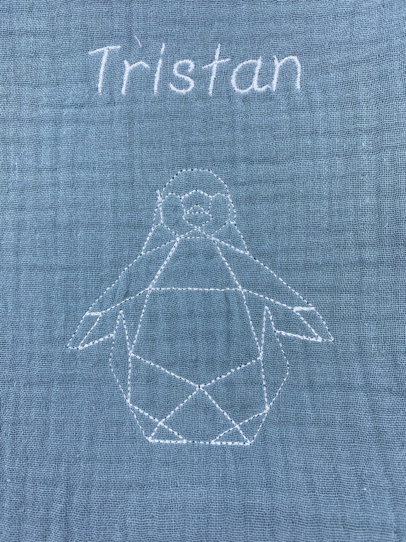 May include: A light blue baby blanket with the name "Tristan" embroidered in white. A white geometric penguin is also embroidered on the blanket.