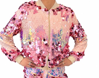 May include: A long-sleeve bomber jacket and matching shorts set covered in iridescent pink and magenta sequins. The jacket features a front zipper closure, a ribbed collar, and ribbed cuffs in a neutral beige tone, creating a cohesive look.