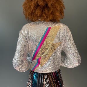 Puede incluir: Una blusa de lentejuelas plateadas con un diseño de rayo dorado, rosa y azul en la espalda.