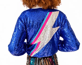 May include: A long-sleeved blue sequin jacket featuring a large silver lightning bolt appliqué on the back, accented with thin diagonal stripes of bright pink and turquoise. The jacket is paired with high-waisted pants featuring vertical stripes.