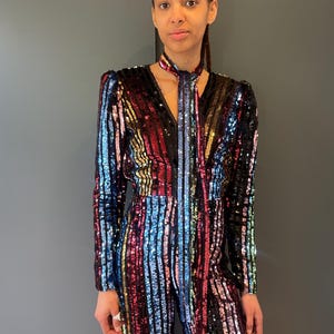 Könnte beinhalten: Ein paillettenbesetzter Overall mit vertikalen Streifen in Regenbogenfarben, darunter Rot, Blau und Gold. Der langärmlige Overall hat einen V-Ausschnitt und eine passende Krawatte. Das Kleidungsstück ist ein einteiliges Outfit.