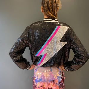 May include: A black sequined bomber jacket with a silver lightning bolt design on the back. The lightning bolt has pink and blue accents. The jacket is paired with pink sequined shorts.