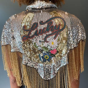 May include: Silver sequined jacket with gold fringe. The back features a horseshoe design with the word "Lucky" embroidered in red, teal, and gold. Floral embroidery adds a pop of color. A stylish, eye-catching piece.