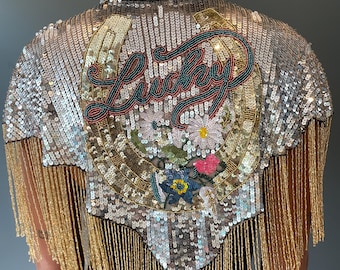 May include: Silver sequined jacket with gold fringe. The back features a horseshoe design with the word "Lucky" embroidered in red, teal, and gold. Floral embroidery adds a pop of color. A stylish, eye-catching piece.