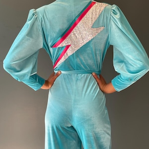May include: A turquoise velvet jumpsuit with long sleeves and wide legs. The back features a sequined lightning bolt design with pink and teal accents. The jumpsuit has a cinched waist and a relaxed fit.