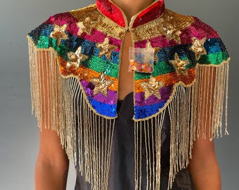 May include: A colorful sequined cape with a rainbow design and gold star accents. The cape features gold fringe and a red velvet collar. The garment is open in the front and appears to be vintage.
