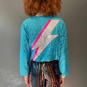 May include: A turquoise sequined long-sleeved top with a silver and pink lightning bolt design on the back. The top is paired with multi-coloured sequined trousers.