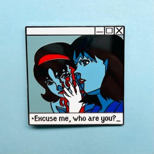 Può includere: Spilla smaltata con un'illustrazione stilizzata di due figure con pelle blu e capelli neri. Una figura sembra toccare il volto dell'altra, con striature rosse che suggeriscono sangue. La spilla include il testo "Excuse me, who are you?"