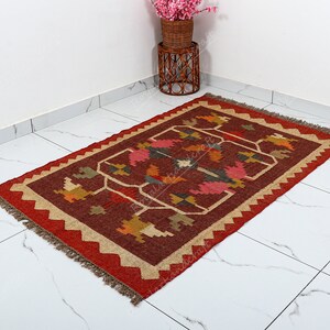 May include: A red, brown, and beige handwoven rug with a geometric pattern. The rug has a fringe border.