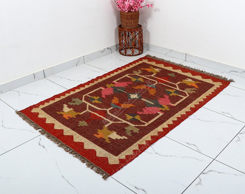 May include: A red, brown, and beige handwoven rug with a geometric pattern. The rug has a fringe border.