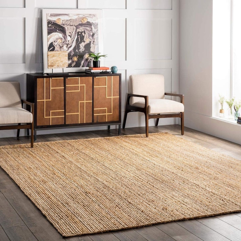 May include: A natural fiber jute rug in a light brown color with a woven texture. The rug is placed in a living room setting with a wooden cabinet and two chairs.
