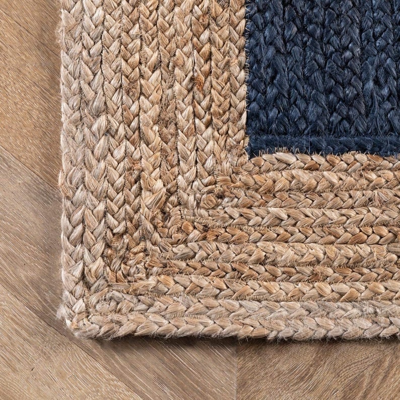 May include: Close-up of a woven jute rug with a blue and natural color pattern. The rug has a braided edge and is made of natural fibers.