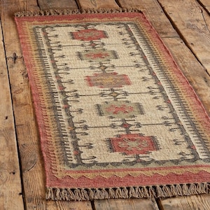May include: Rectangular jute rug with a geometric pattern in red, beige, and grey. The rug has a fringed edge and is placed on a wooden floor. The design features repeating rectangular and diamond shapes.