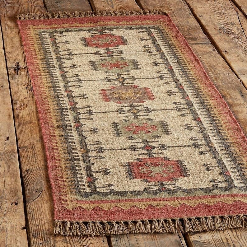 May include: Rectangular jute rug with a geometric pattern in red, beige, and grey. The rug has a fringed edge and is placed on a wooden floor. The design features repeating rectangular and diamond shapes.
