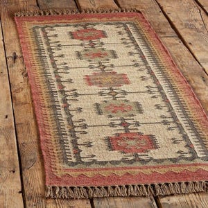 May include: Rectangular beige rug with a geometric pattern in red, brown, and green. The rug has a fringed edge and a border with a repeating design. The rug is made of a woven material and is placed on a wooden floor.