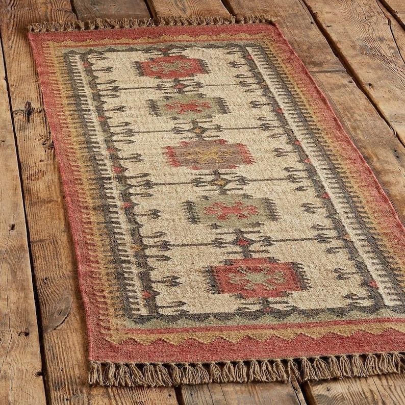 May include: Rectangular beige rug with a geometric pattern in red, brown, and green. The rug has a fringed edge and a border with a repeating design. The rug is made of a woven material and is placed on a wooden floor.