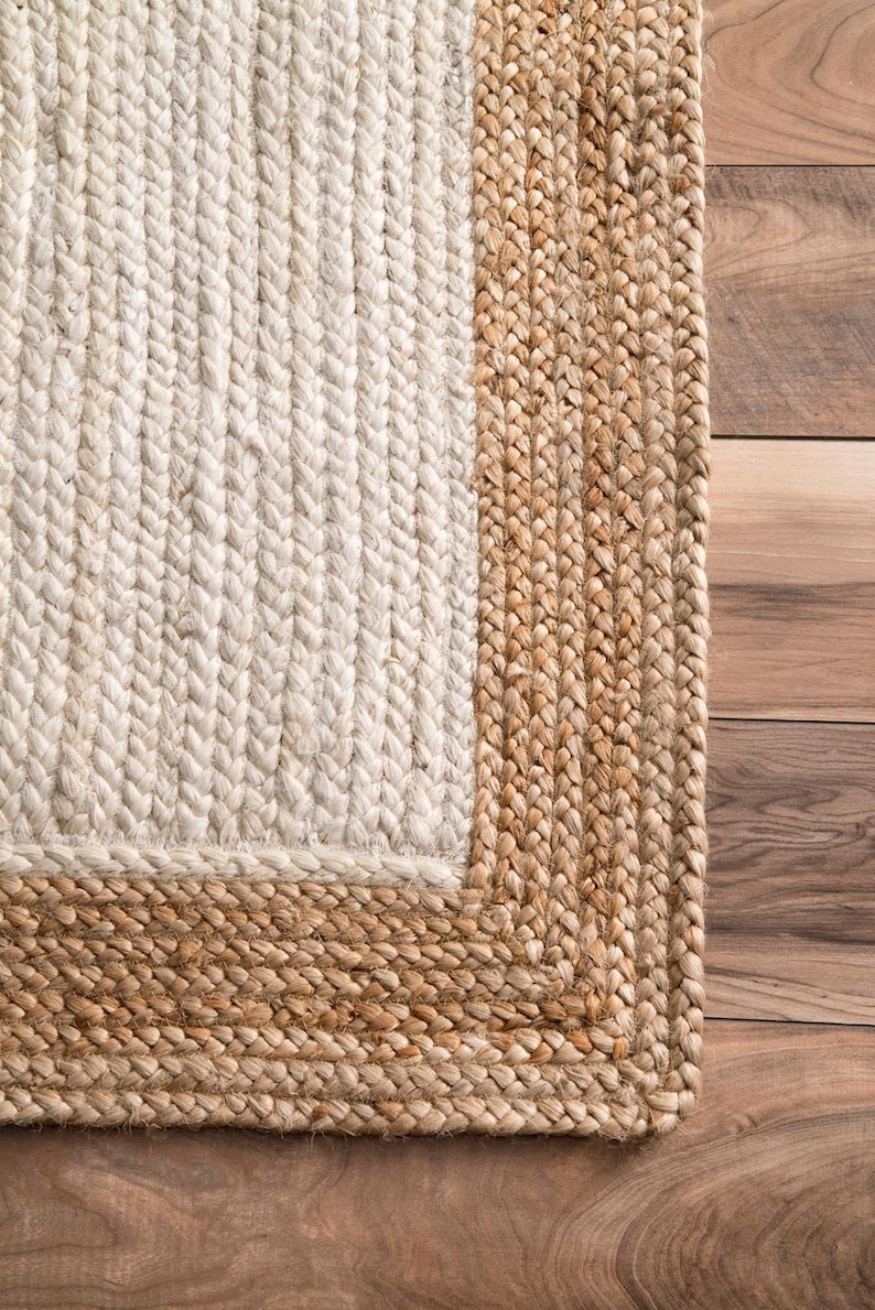 May include: A natural fiber rug with a white center and a brown border. The rug is made of woven jute and has a textured surface.