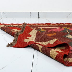 May include: A red, brown, and beige handwoven rug with a geometric pattern. The rug is folded in half and has fringe on the edges.
