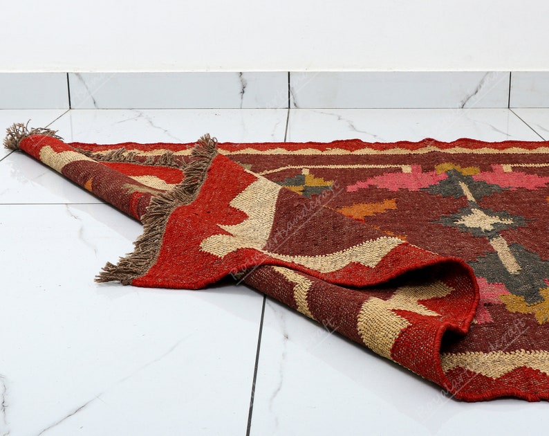 May include: A red, brown, and beige handwoven rug with a geometric pattern. The rug is folded in half and has fringe on the edges.