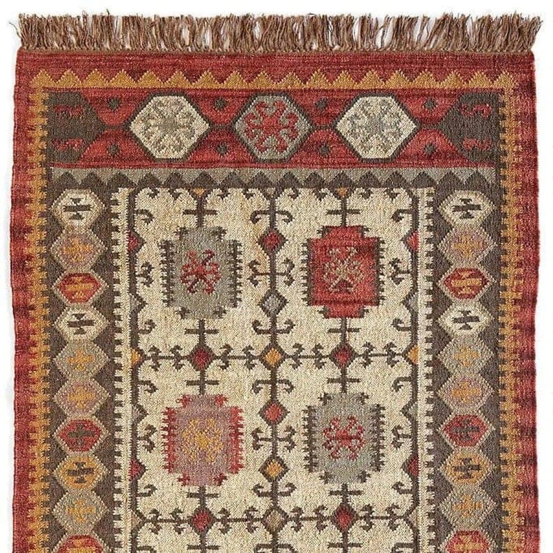 May include: Close-up of a rectangular rug with a detailed geometric pattern. The rug features a cream-coloured central field with repeating motifs in red, brown, grey, and yellow. A red border with a fringe edge frames the design, showcasing a traditional style.