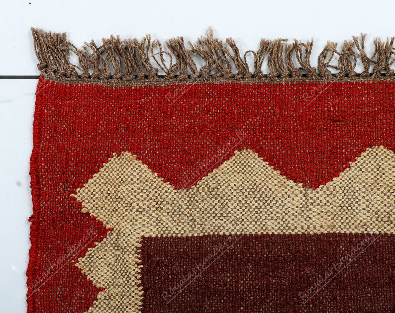 May include: Close-up of a red, brown, and beige woven rug with a geometric pattern. The rug has a fringe border.
