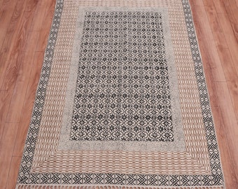 Puede incluir: Una alfombra blanca y negra con estampado, borde marrón y flecos. La alfombra tiene un diseño geométrico con un motivo floral.