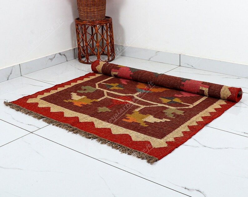 May include: A red, brown, and beige handwoven rug with a geometric pattern. The rug has a fringe border and is rolled up on a white tiled floor.
