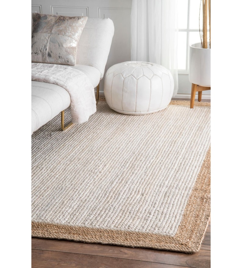 May include: A white and beige jute rug with a natural jute border. The rug is shown in a living room setting with a white sofa and a white pouf.