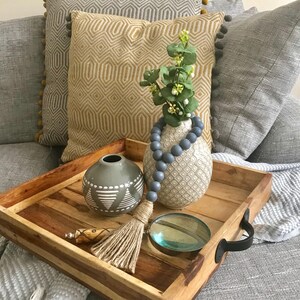 May include: A wooden tray with a gray and white patterned vase with blue beads, a gray vase with a white geometric design, a natural fiber tassel, and a magnifying glass.