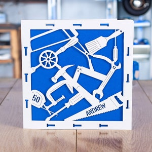 May include: A white paper cut-out card with a blue background featuring various tools, including a hammer, saw, wrench, and screwdriver. The card says "Andrew" and "50" in the design.