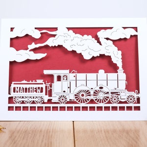 May include: White paper cut-out card with a red background. The card features a steam train with the name "MATTHEW" on the side. The train is surrounded by white clouds and smoke.
