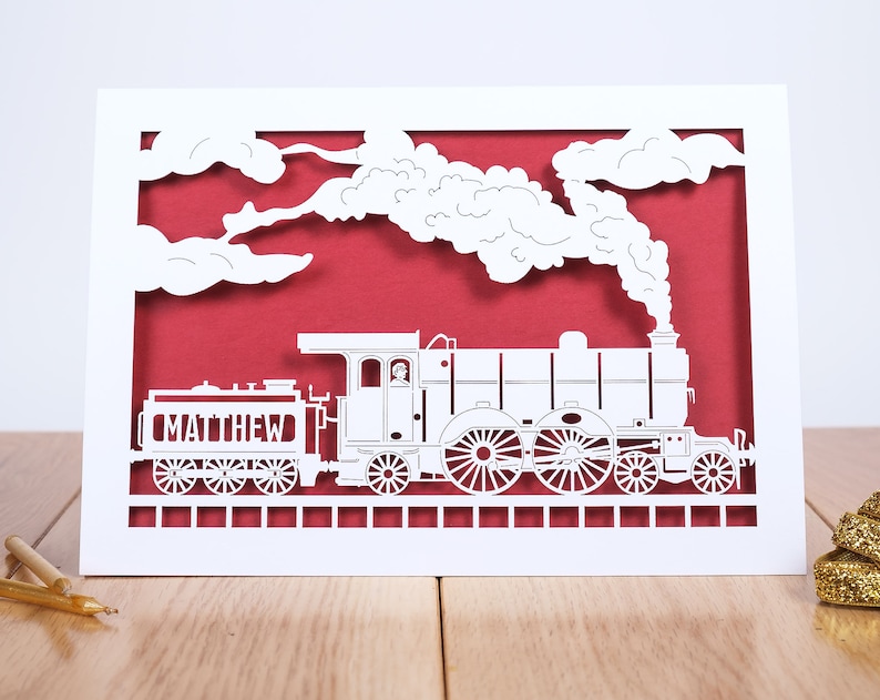 May include: White paper cut-out card with a red background. The card features a steam train with the name "MATTHEW" on the side. The train is surrounded by white clouds and smoke.