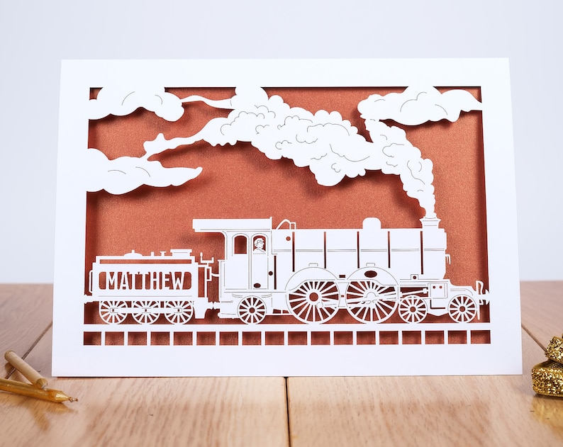 May include: White paper card with a cut-out design of a steam train with smoke and clouds. The train is labeled "MATTHEW".