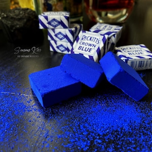 May include: Three blue blocks of Reckitt's Crown Blue laundry detergent, with the packaging visible in the background. The blocks are on a black surface with blue powder scattered around them.