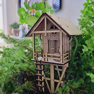 Puede incluir: Un modelo de madera de una pequeña casa con un techo marrón y una ventana. La casa está sobre pilotes y tiene una escalera que lleva a la puerta principal. La casa está rodeada de plantas verdes.