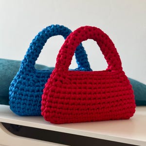 May include: Two crocheted handbags, one red and one blue. The bags have a simple, rectangular shape with a rounded top and a single handle.